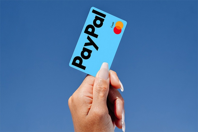 A hand holding a light blue PayPal credit card featuring the Mastercard logo, set against a clear blue sky.